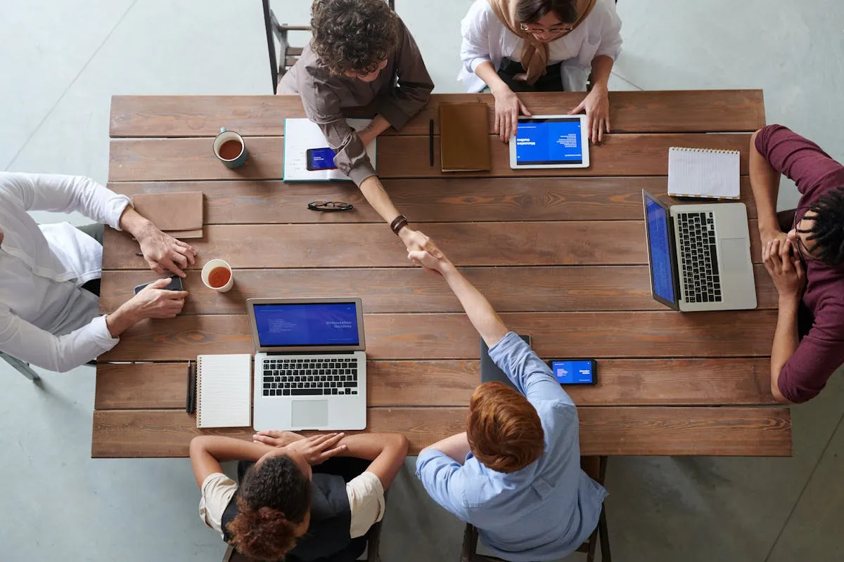 Team meeting around a table with laptops, shaking hands on a partnership